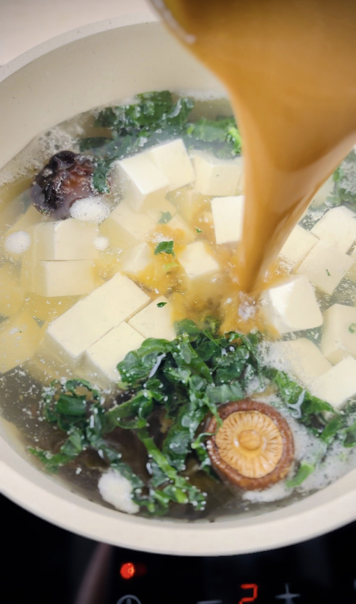 Miso and water mixture being poured into saucepan full of broth and tofu cubes.