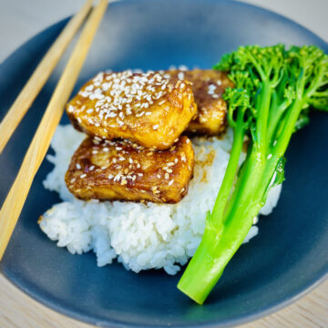 Three tofu steaks, sprinkled with sesame seeds, resting on top of a bowl filled with steamed white rice and broccolini.