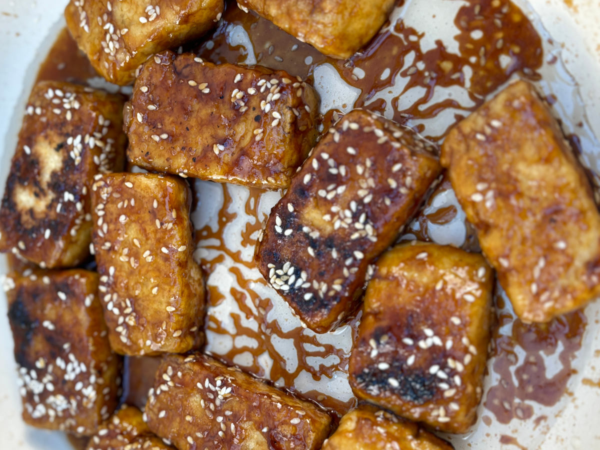 A dozen tofu steaks frying in a pan, almost fully done, a lovely deep brown color and covered in sticky teriyaki sauce and white sesame seeds.