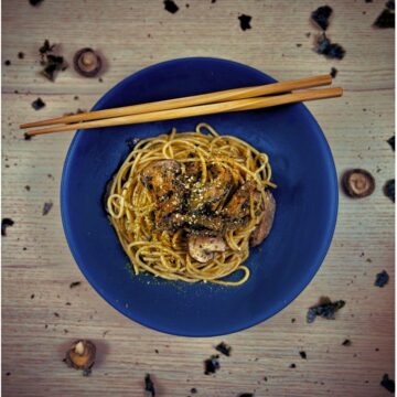 A ceramic bowl, with chopsticks resting on the edge, containing spaghetti topped with mushrooms, seaweed flakes and red Japanese spice powder.