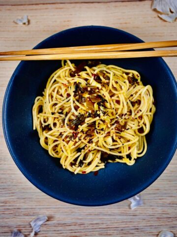 A dark blue pasta bowl containing cooked spaghetti with takana, chilli and seasoning on top; plus a pair of chopsticks rested on the edge of the bowl.