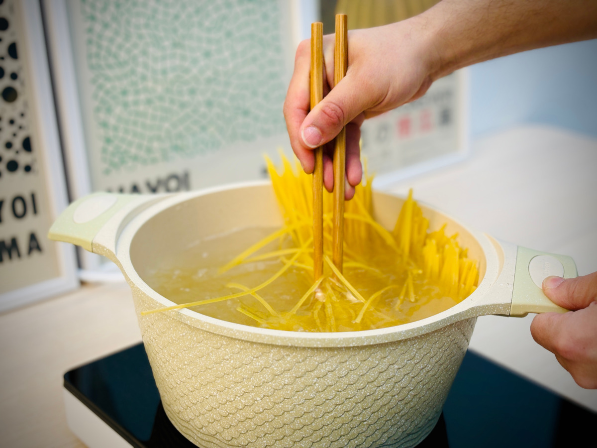 Pot of water on an electric stove containing water and dried spaghetti; a hand holding a pair of wooden chopsticks is picking up some of the slightly limp spaghetti pieces in the water.