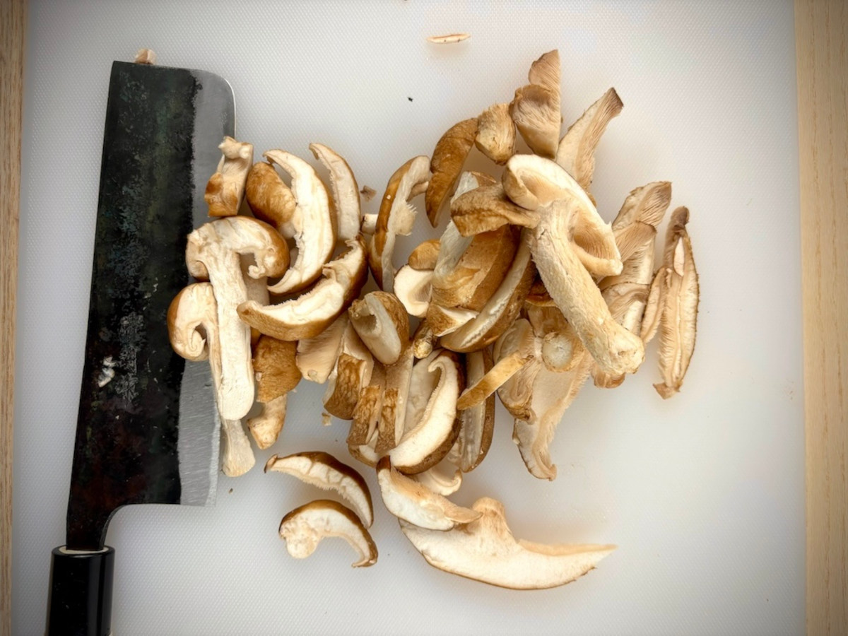 A white chopping board with a pile of 1cm thick sliced mushrooms sitting in the middle, and a large Japanese chopping knife resting on the left side.