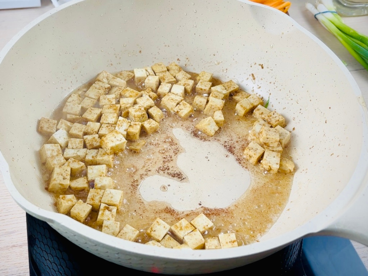 White cubes of tofu in a frying pan with a clear oily liquid and brown powder sprinkled on top.