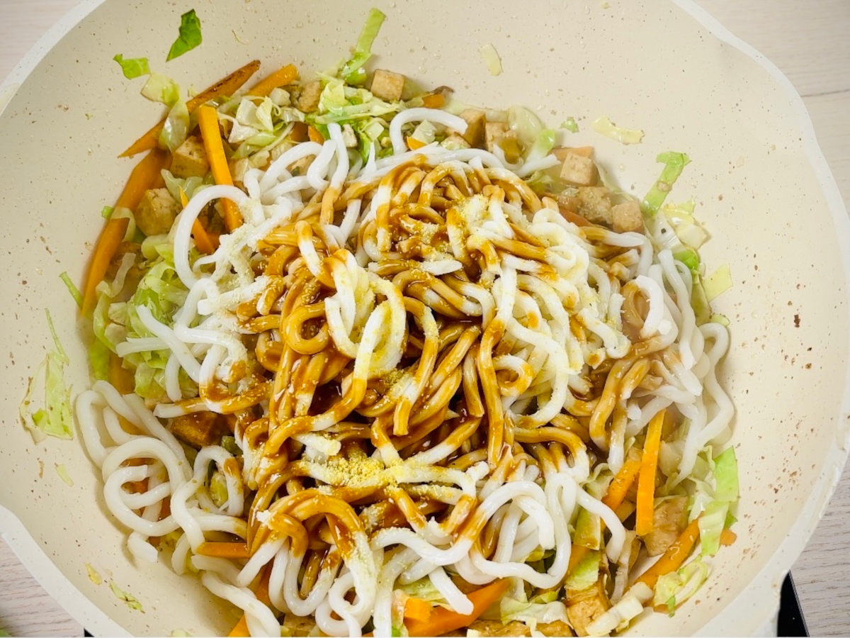 White pan containing chopped vegetables, topped with white noodles, brown sauce and yellow powder.