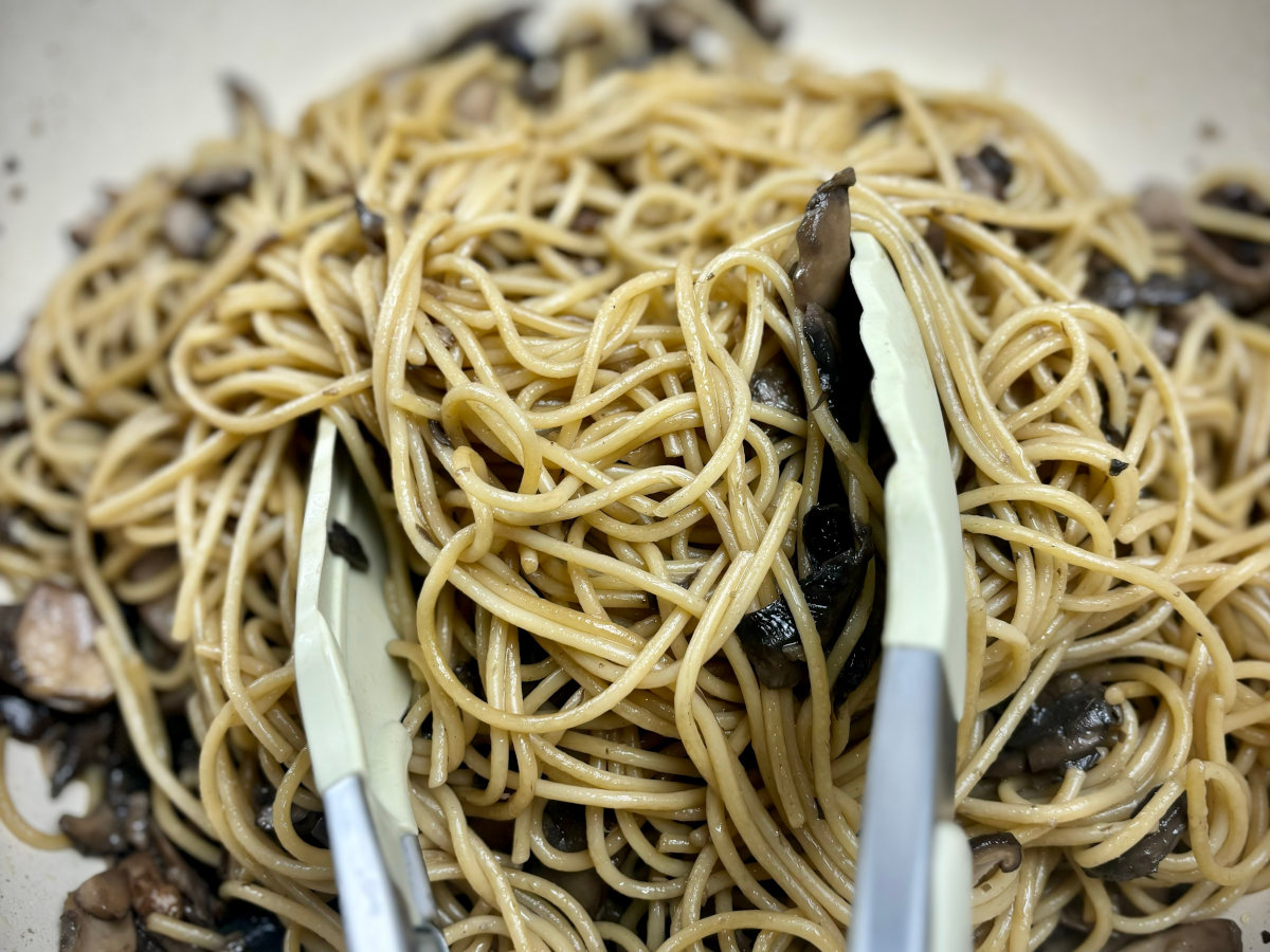 Pair of tongs handling cooked spaghetti with mushrooms combined through it.