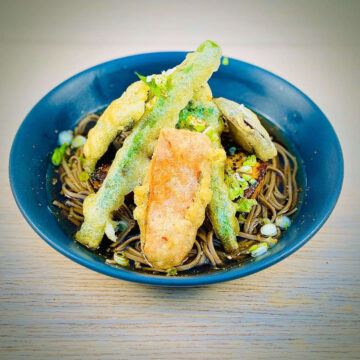 Bowl of noodle soup topped with fried green and orange vegetables covered in crispy batter, garnished with fresh green and white spring onions.