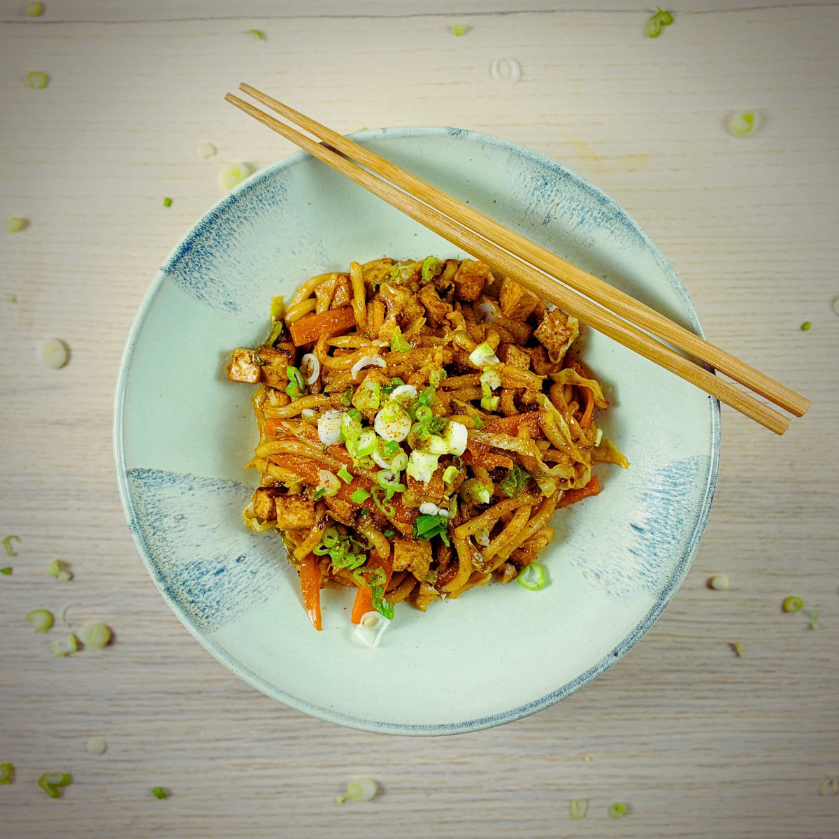 Bowl of cooked noodles with fried tofu, chopped carrots and spring onions on top, with a pair of chopsticks resting on the edge of the bowl.