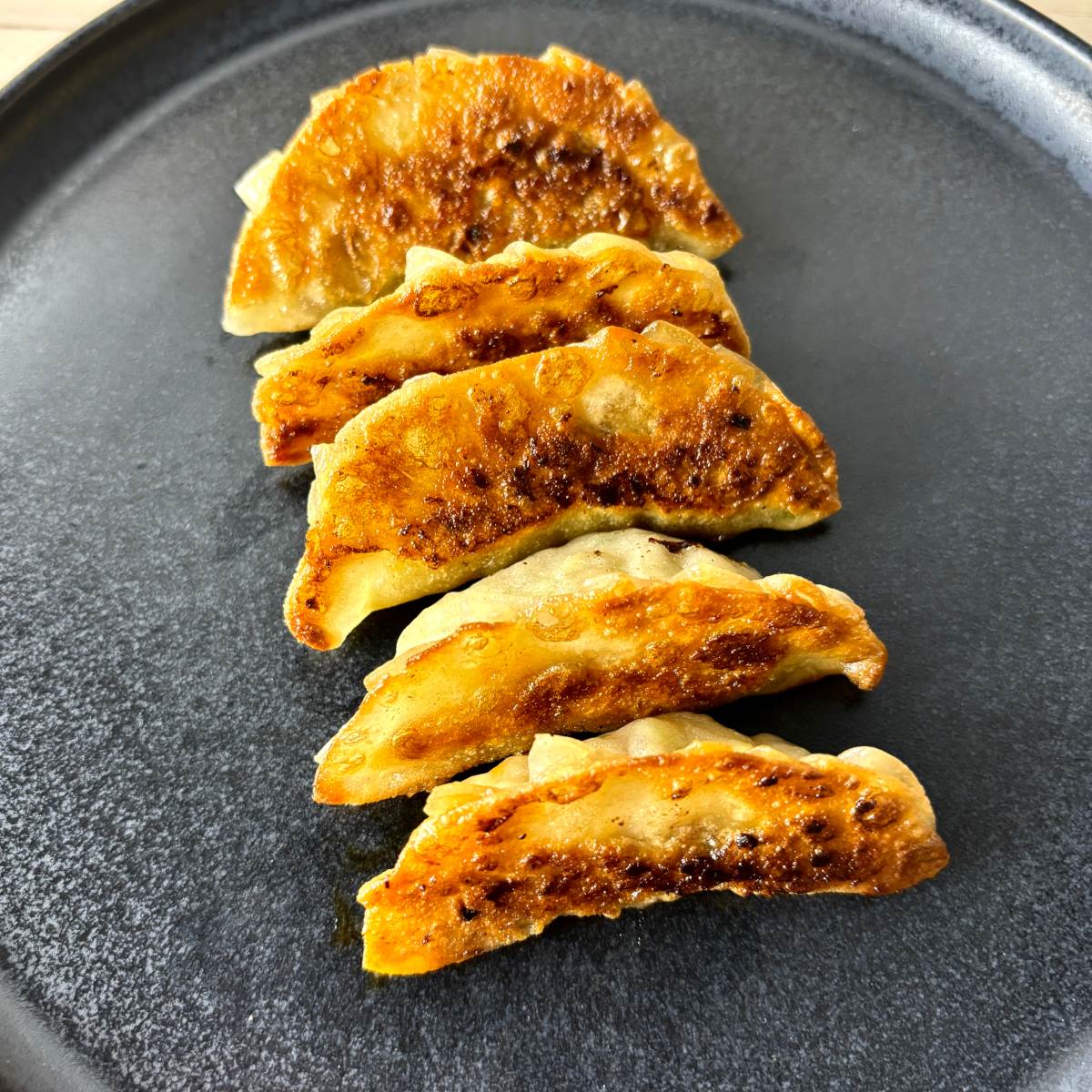 Five crispy-fried gyoza dumplings on a dark charcoal coloured ceramic plate.
