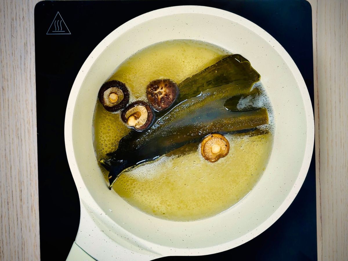 Saucepan on an electric stovetop containing boiling water, four dried shitake mushrooms and two strips of kombu seaweed.