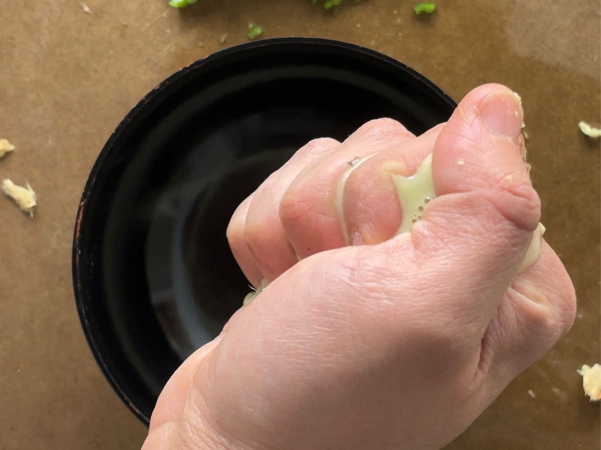 A human hand squeezing ginger pulp over a small black bowl. The pale yellow-coloured juice is coming out to spill over, into the bowl.