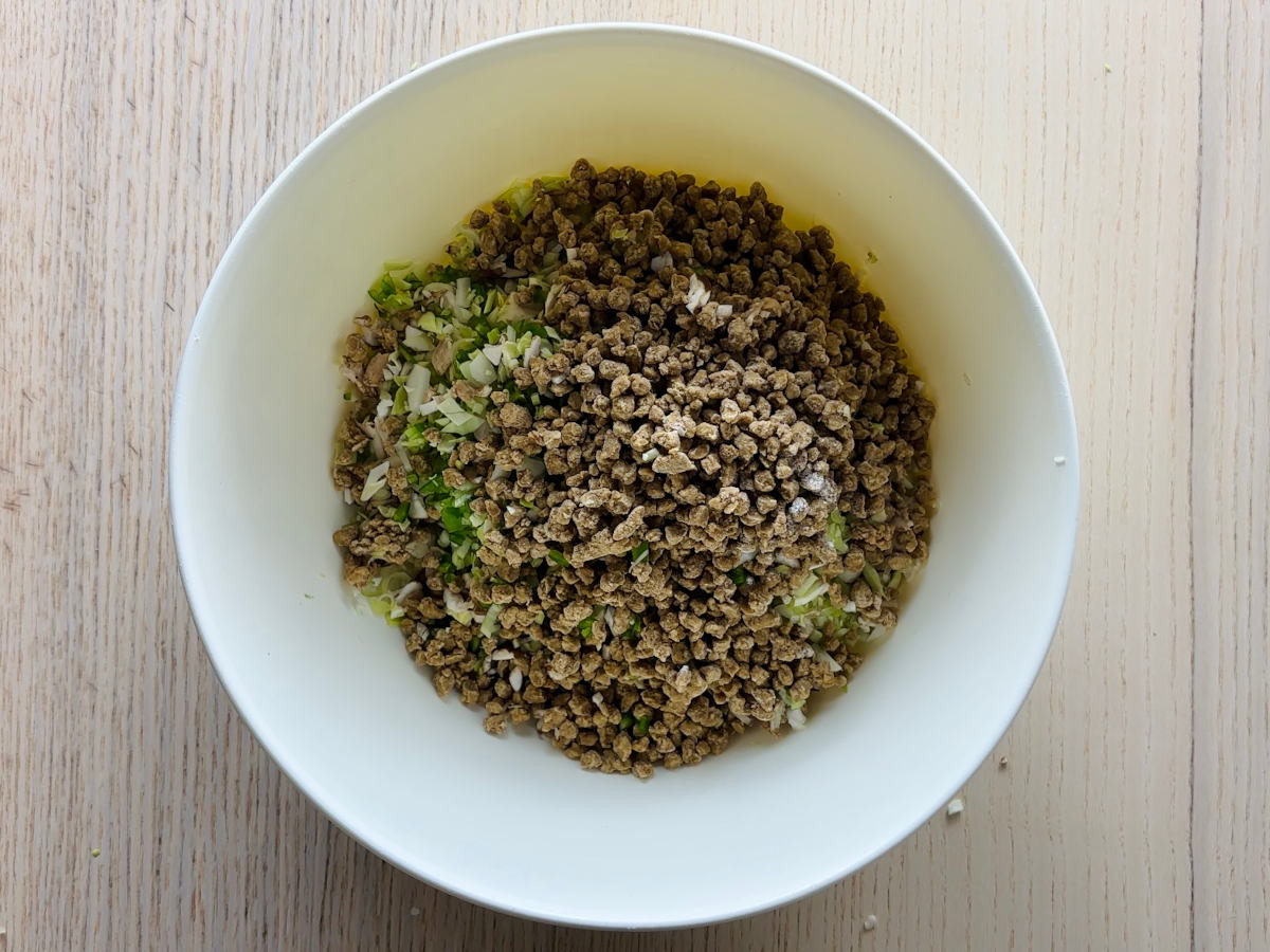 A large white mixing bowl, about half full, containing a mixture of finely chopped green, white and brown vegetables and mushrooms, and a pile of frozen brown granules of soya mince on top.