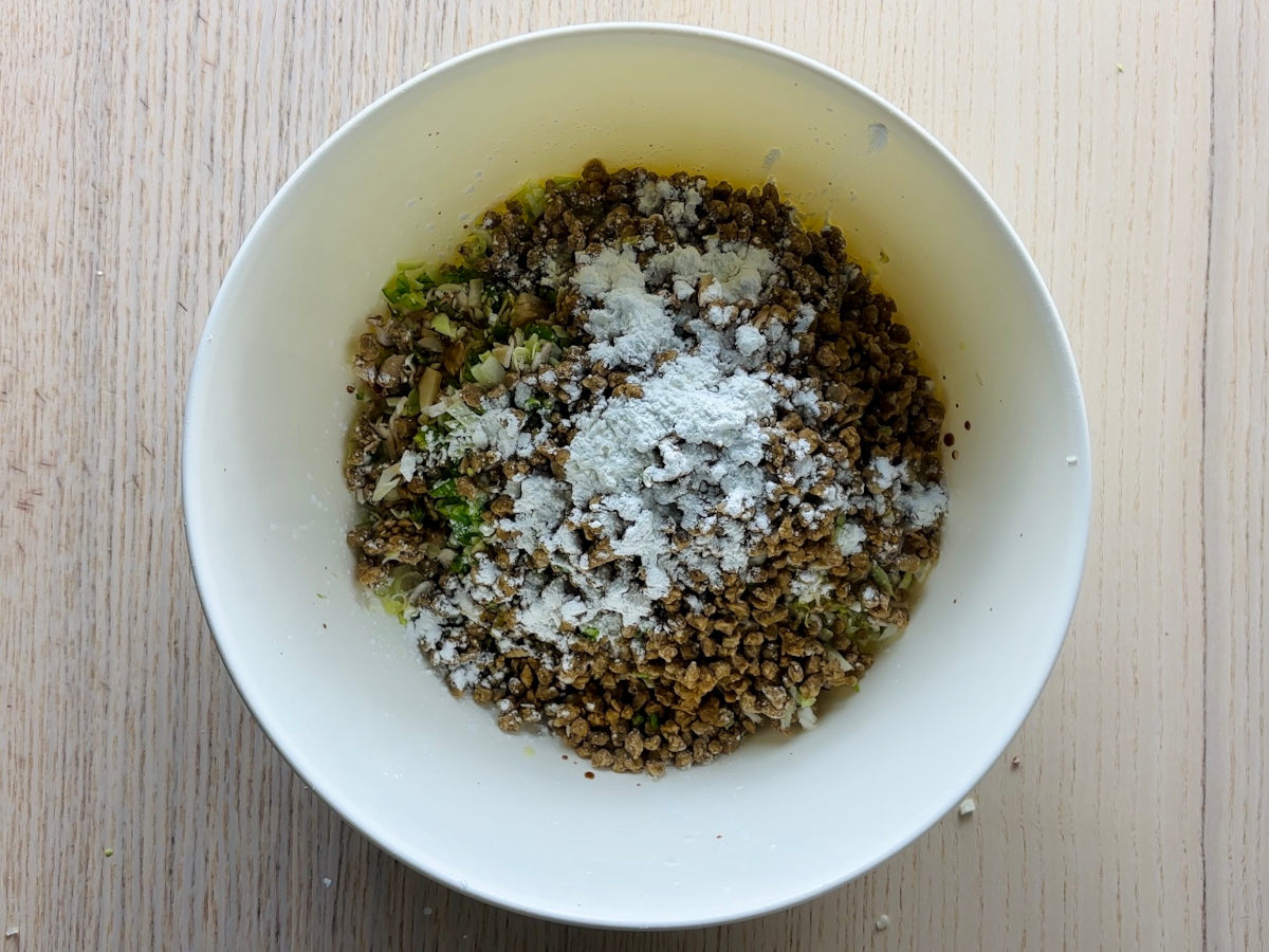 A large white mixing bowl containing finely diced vegetables, mushrooms and brown granules of frozen soya mince, with a small pile of seasonings and white cornflour on top, with a few splashes of dark brow soya sauce around the edge of the bowl.
