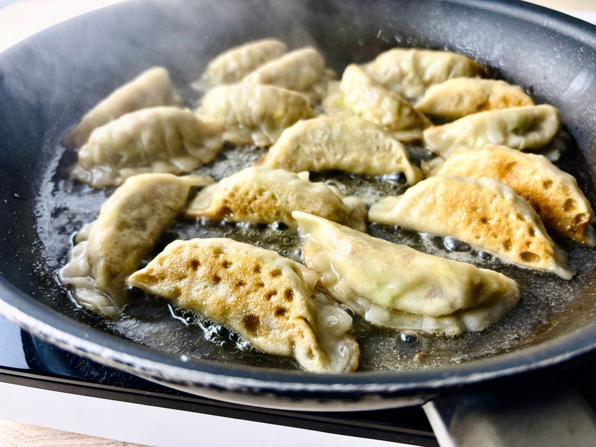 A medium frying pan containing about 16 gyoza dumplings, that appear to be sizzling and frying in a shallow pool of bubbling clear liquid, with steam coming off the top.