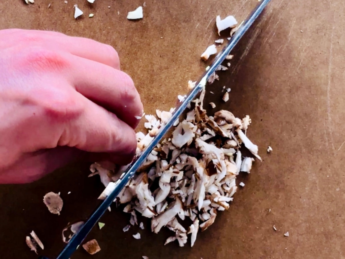 A human hand and knife holding and finely chopping mushroom slices on a brown chopping board.