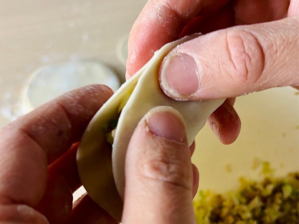 A close up of a person's left and right forefingers and thumbs folding a gyoza wrapper in half so that the opposite edges meet, while the middle of the gyoza wrapper contains filling, creating a little parcel.