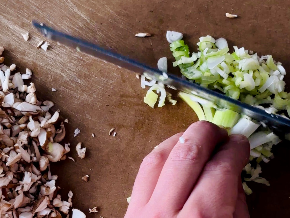 A human hand holding spring onion stalks next to a knife in process of slicing them thinly. A pile of finely diced mushrooms is sitting to the left of the hand on teh same chopping board.