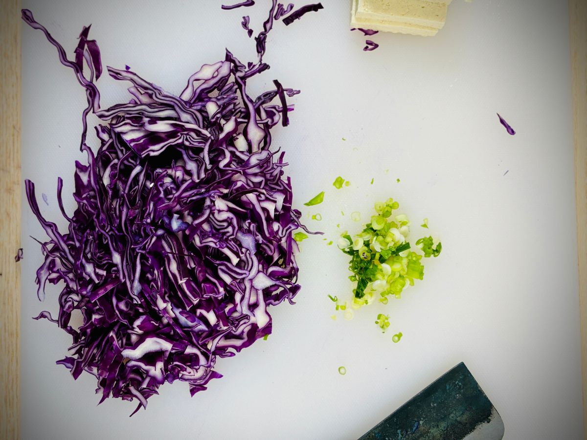 A handful of finely chopped red cabbage next to a small pile of finely chopped spring onions on a white chopping board.