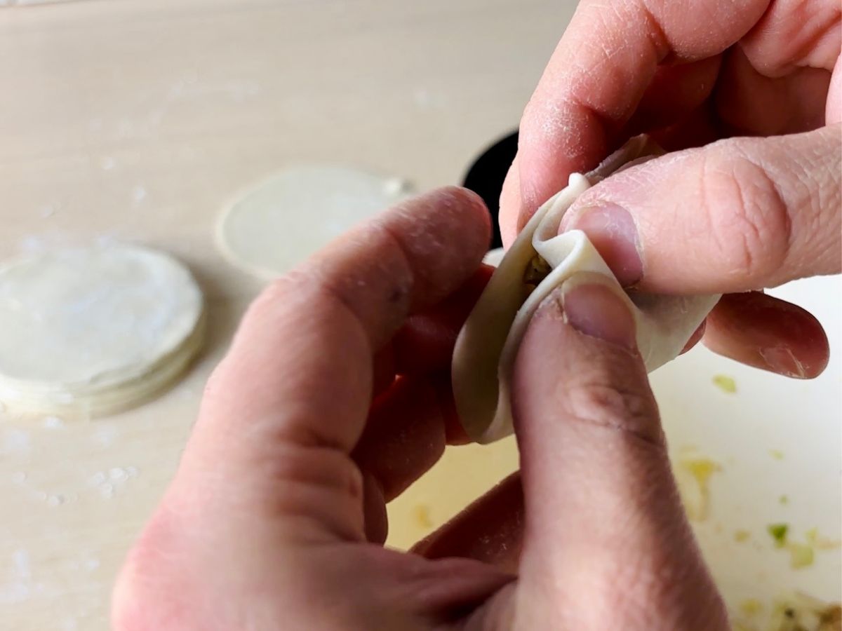 A person's left and right hands holding an uncooked gyoza dumpling, creating a pleat or fold on one of the edges.