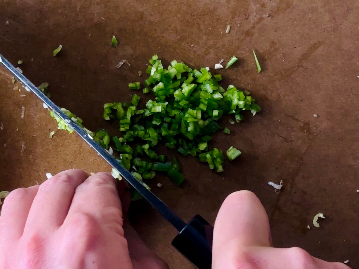 A hand and knife holding and finely chopping fresh green chilli on a brown chopping board.