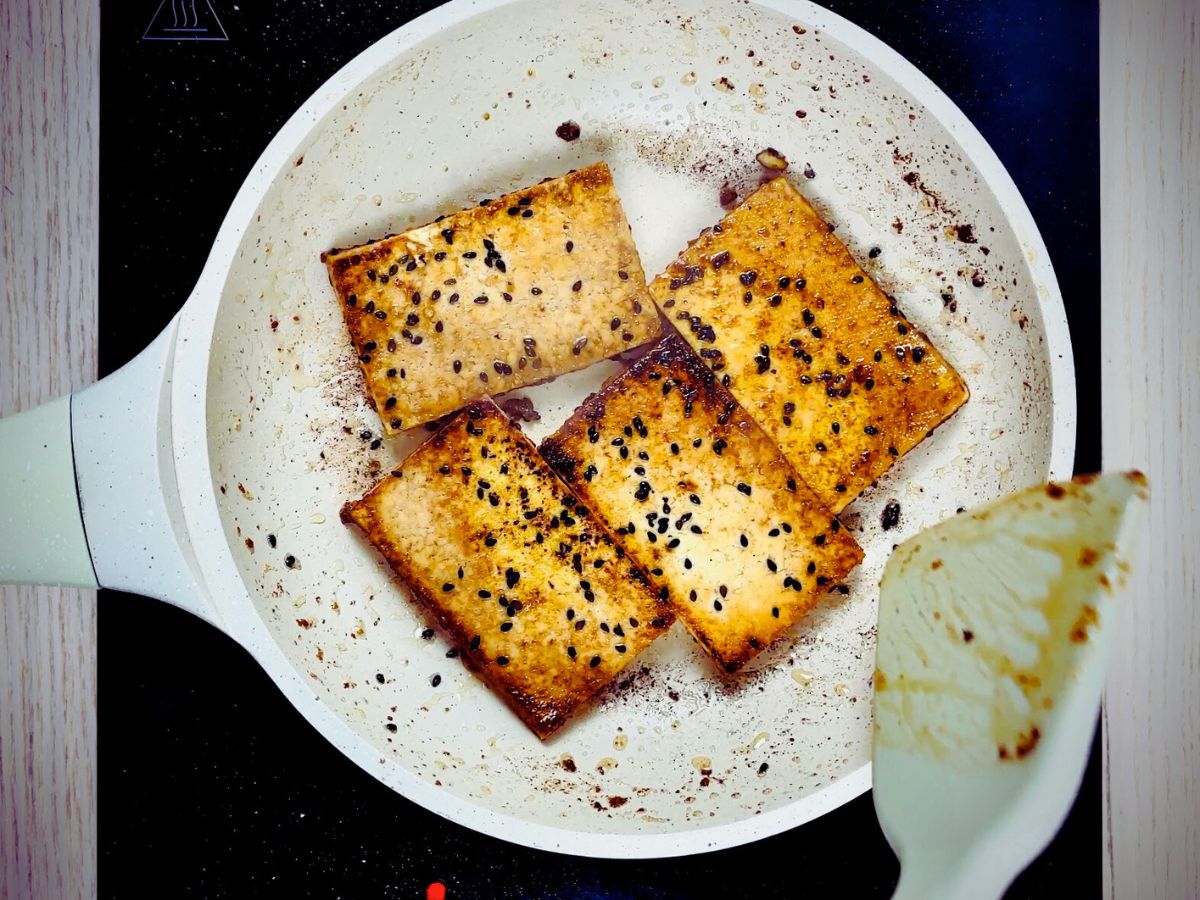 Frying pan on an electric stovetop containing four browned tofu steaks lightly coated with black seeds.