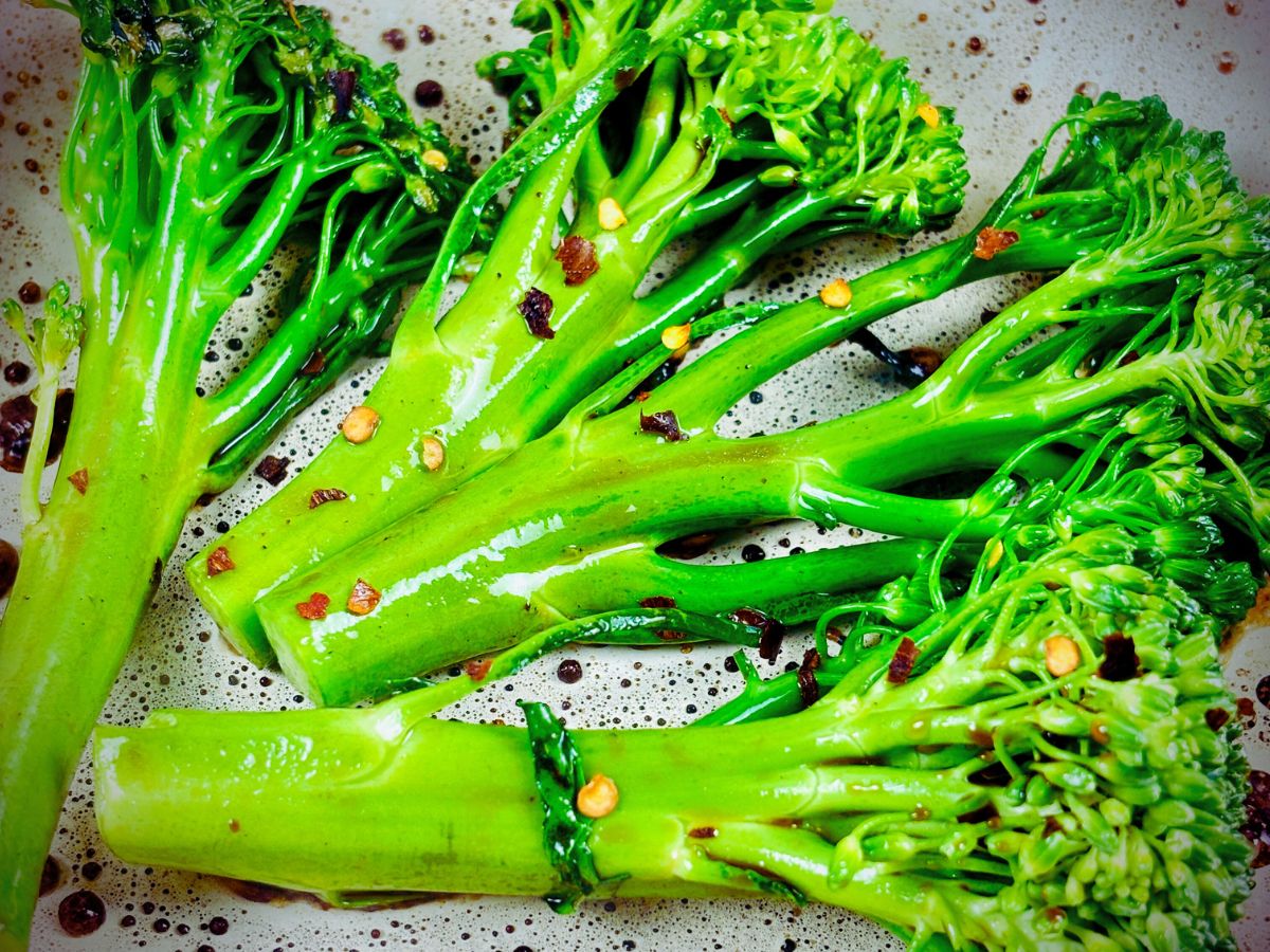 Close-up photo of four cooked broccolini stems coated in oil and red and yellow flakes (dried crushed chilli).