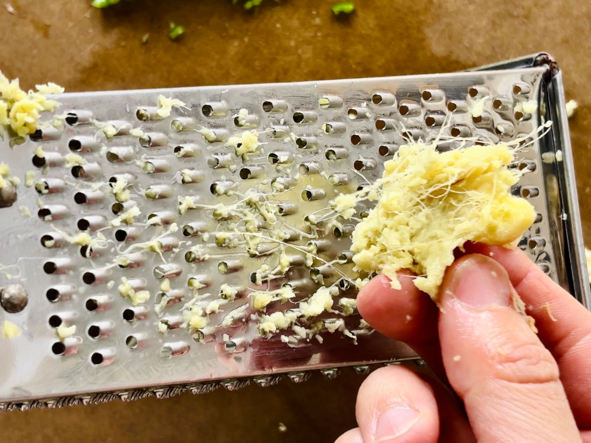 A hand holding a piece of ginger next to a fine steel metal grater.