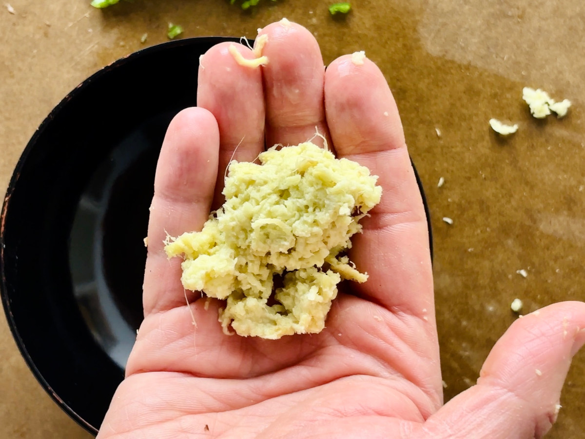 A person's open hand, containing a cluster of freshly grated ginger pulp, with a small black bowl in the background.