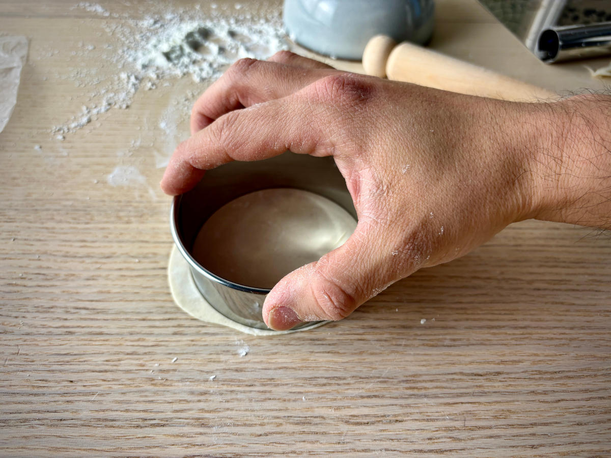 A hand holding a round cookie cutter over some flat, rolled-out dough, ready to cut it into a small circle shape.