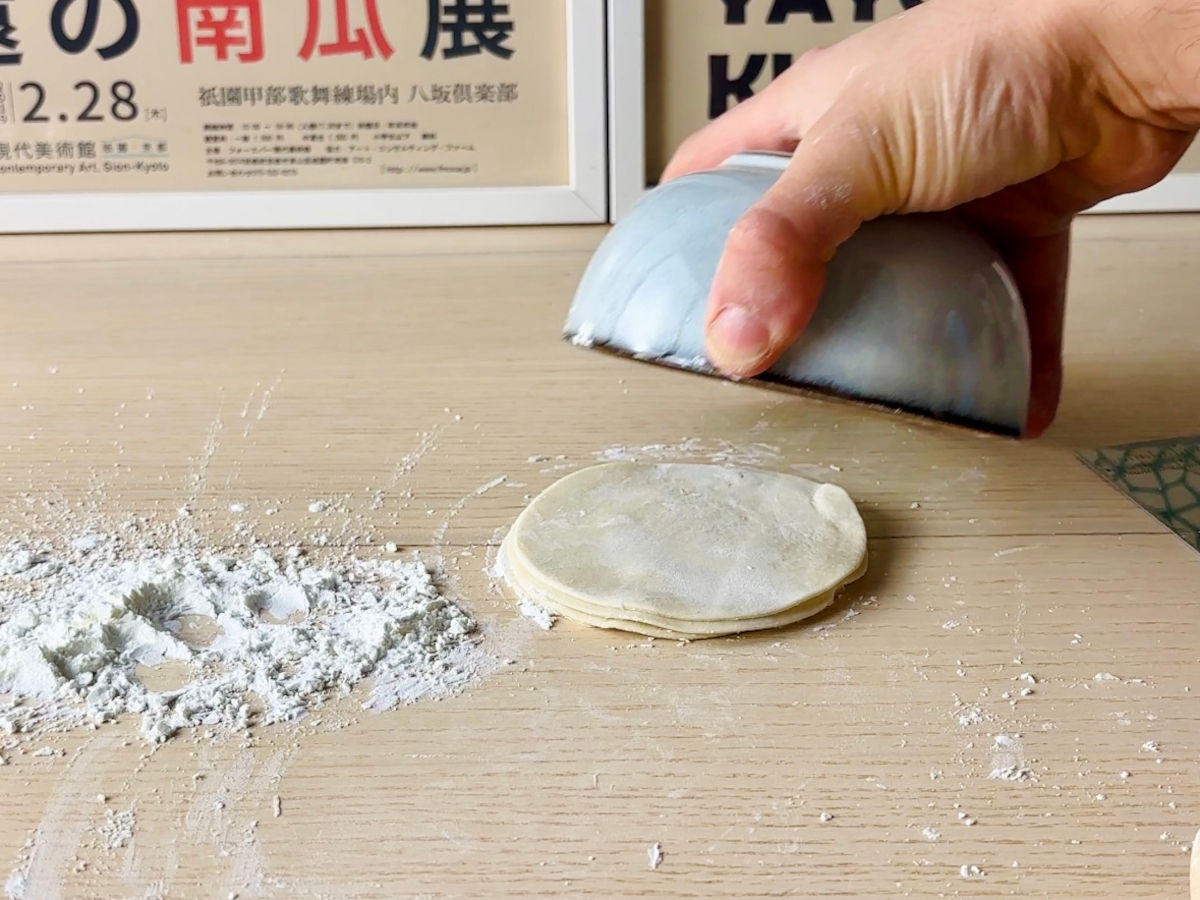 A small pile of fresh gyoza wrappers, stacked on a table, next to some sprinklings of white flour. A hand is holding a small bowl over the pile of wrappers to cover them so they stay fresh.