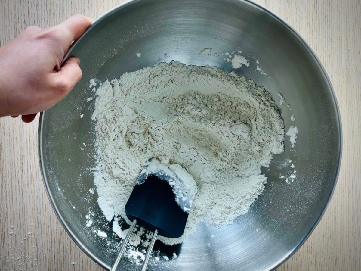 A large metal bowl containing a black spatula that is mixing some white flour and salt with a small amount of water.