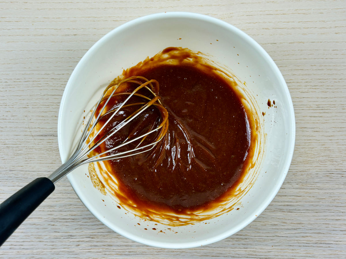 A large mixing bowl containing a deep-brown shiny coloured sauce being whisked smooth, to make a vegan Japanese tonkatsu sauce.