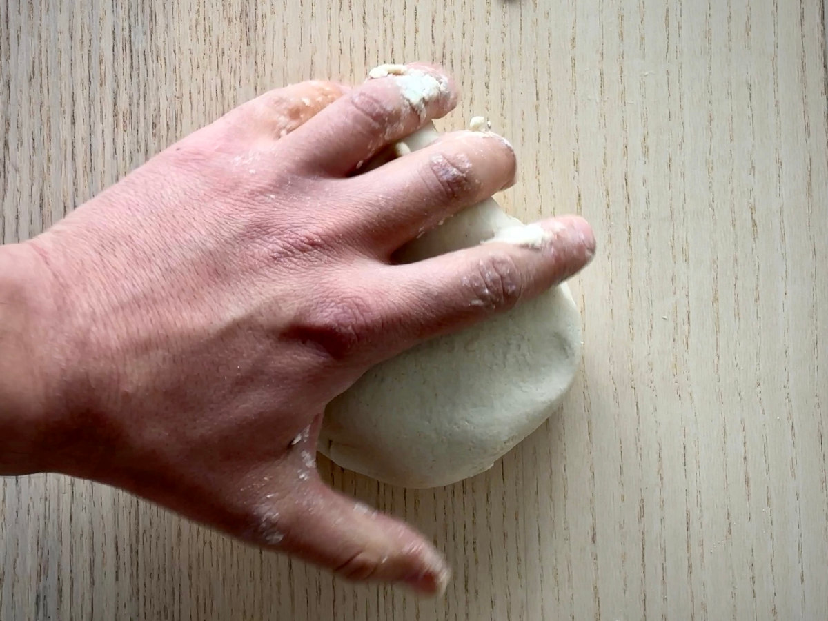 A hand kneading a round ball of dough on a wooden tabletop surface.