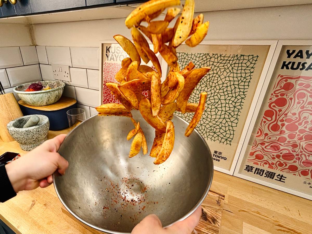 An action shot of someone tossing a serving of vegan Japanese togarashi fries, seasoned with shichimi togarashi spices, in a large metal bowl.