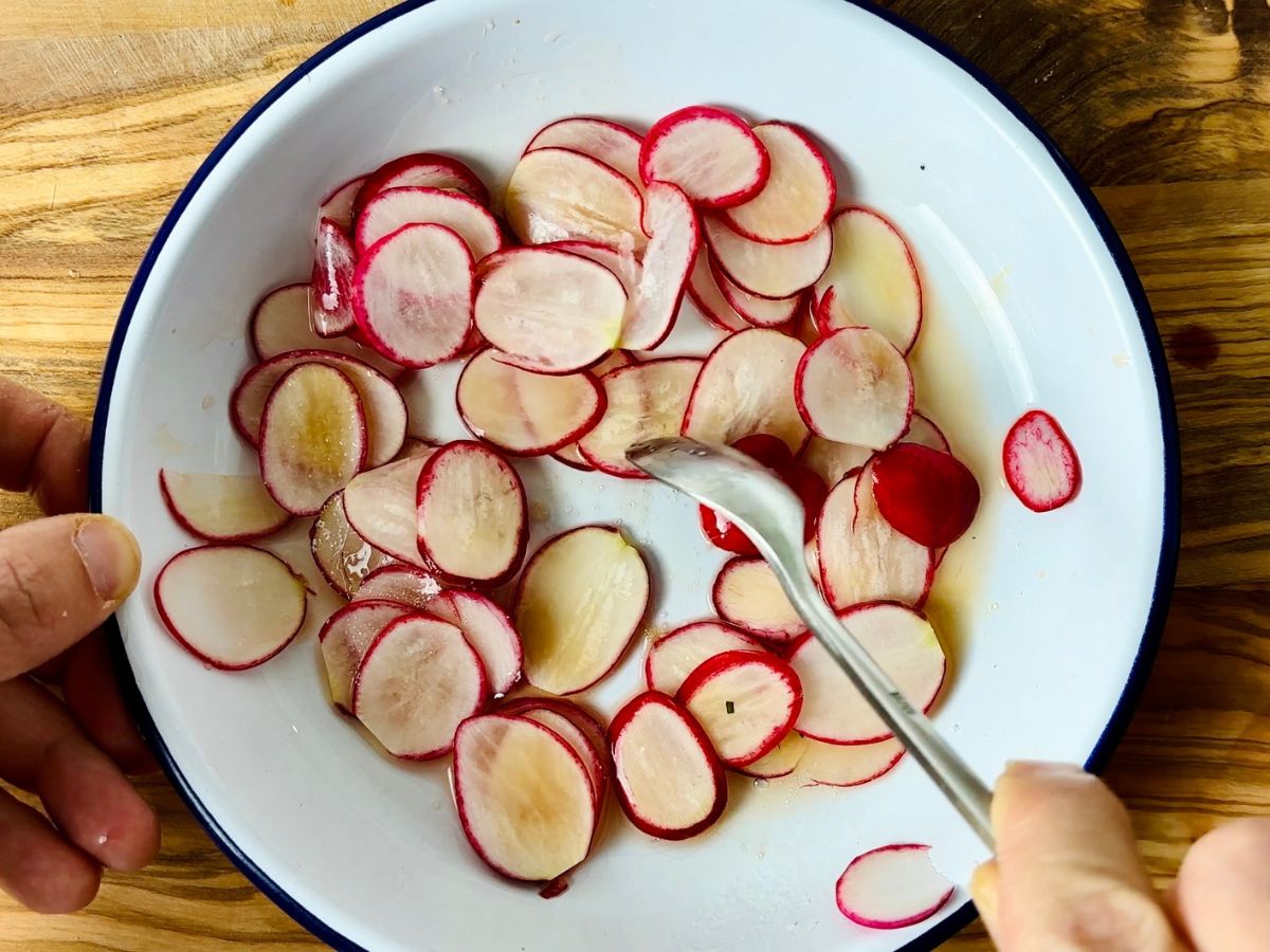 An action shot of thinly sliced pink and white radishes being stirred in some yellowish liquid in a shallow white dish.