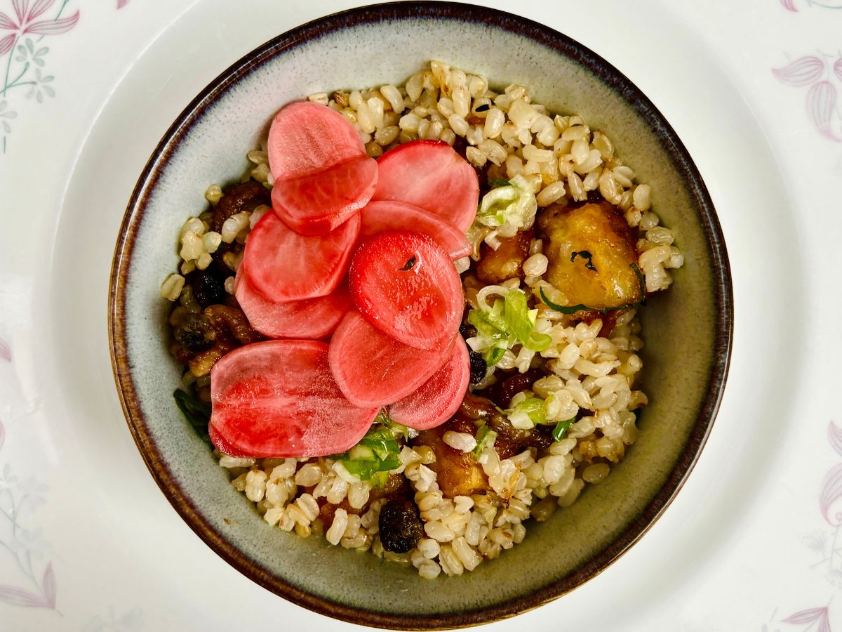 A bowl of rice, tofu and vegetables, topped with sliced pink radish pickles.