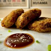 Three golden brown and crispy deep fried Japanese croquettes (korokke), sprinkled with thinly sliced spring onions, next to a circle of brown tonkatsu sauce sprinkled with white sesame seeds on a white marbled serving plate.
