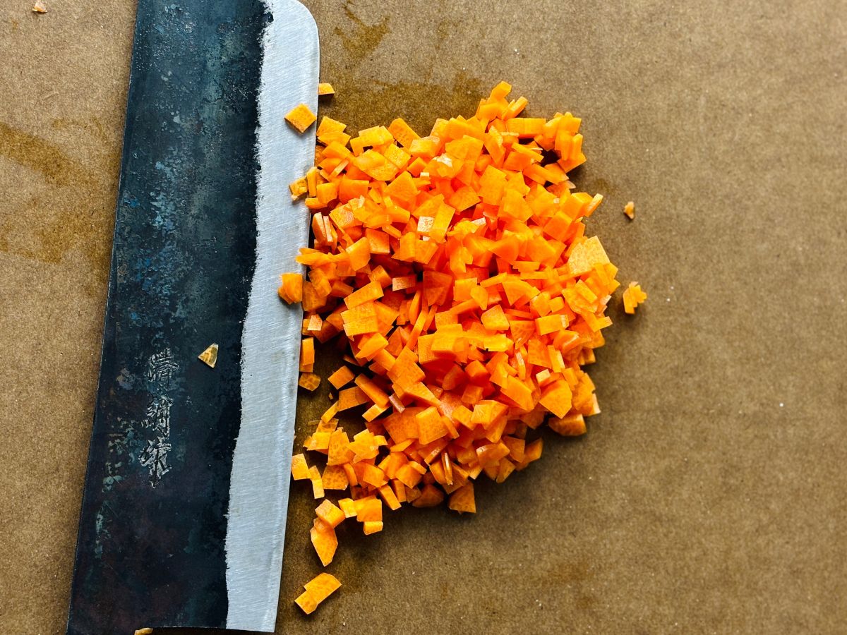 A chopping board with a pile of finely chopped raw carrots on it next to a large kitchen knife.