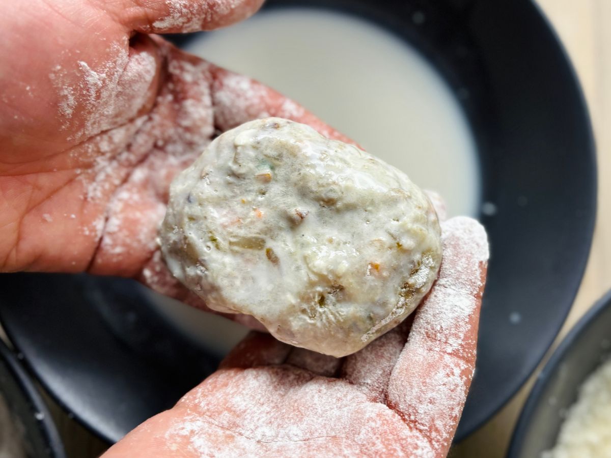 An oval-shaped piece of potato mixture for Japanese croquettes (korokke) that has just been dipped in white liquid milk. A person's flour-covered hands are holding the croquette just above the bowl of milk.