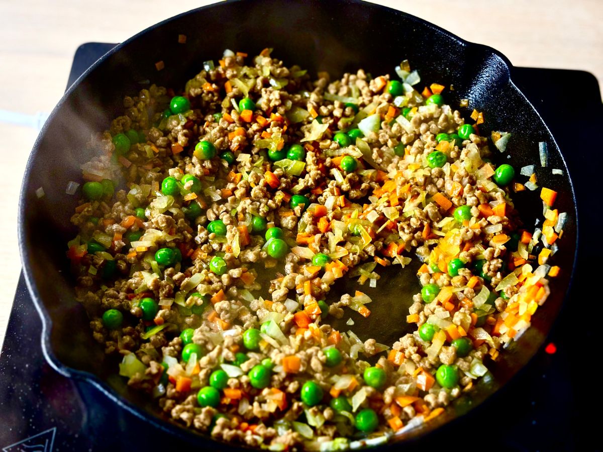 A frying pan on heat, full of a mixture of finely chopped vegetables and green peas with steam coming off the top.