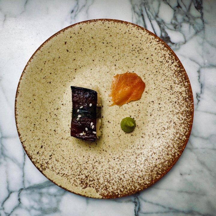 one piece of vegan eel sushi on a round plate next to a petal of sushi giner and a dollop of wasabi