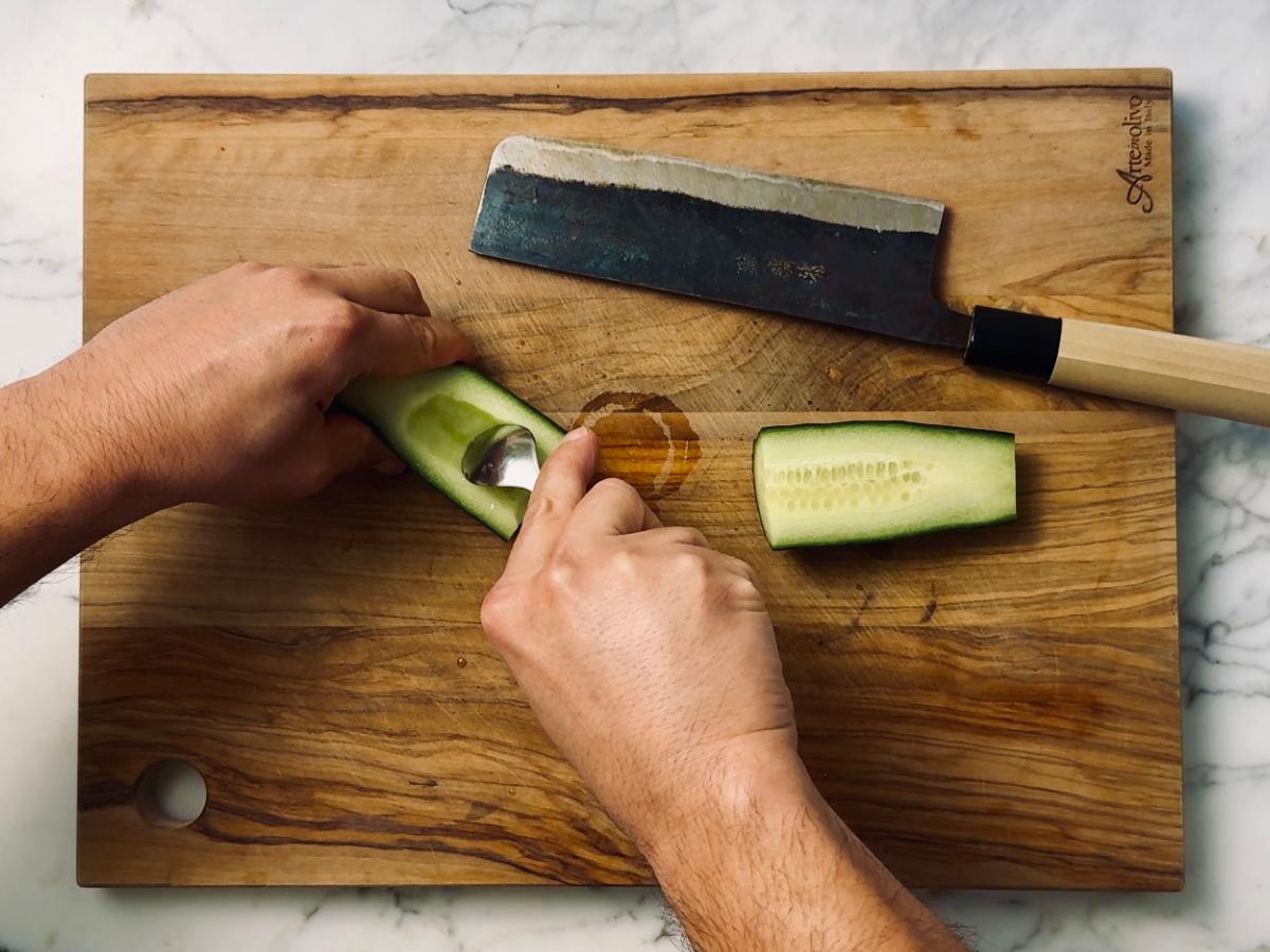 A cucumber half on a chopping board with a teaspoon scooping out the seeds; the other half rests beside it.