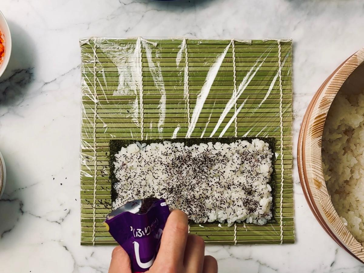 Sushi rice evenly spread across the nori sheet, with shiso furikake being sprinkled lightly over the top. The cling film and bamboo mat are visible beneath the nori.