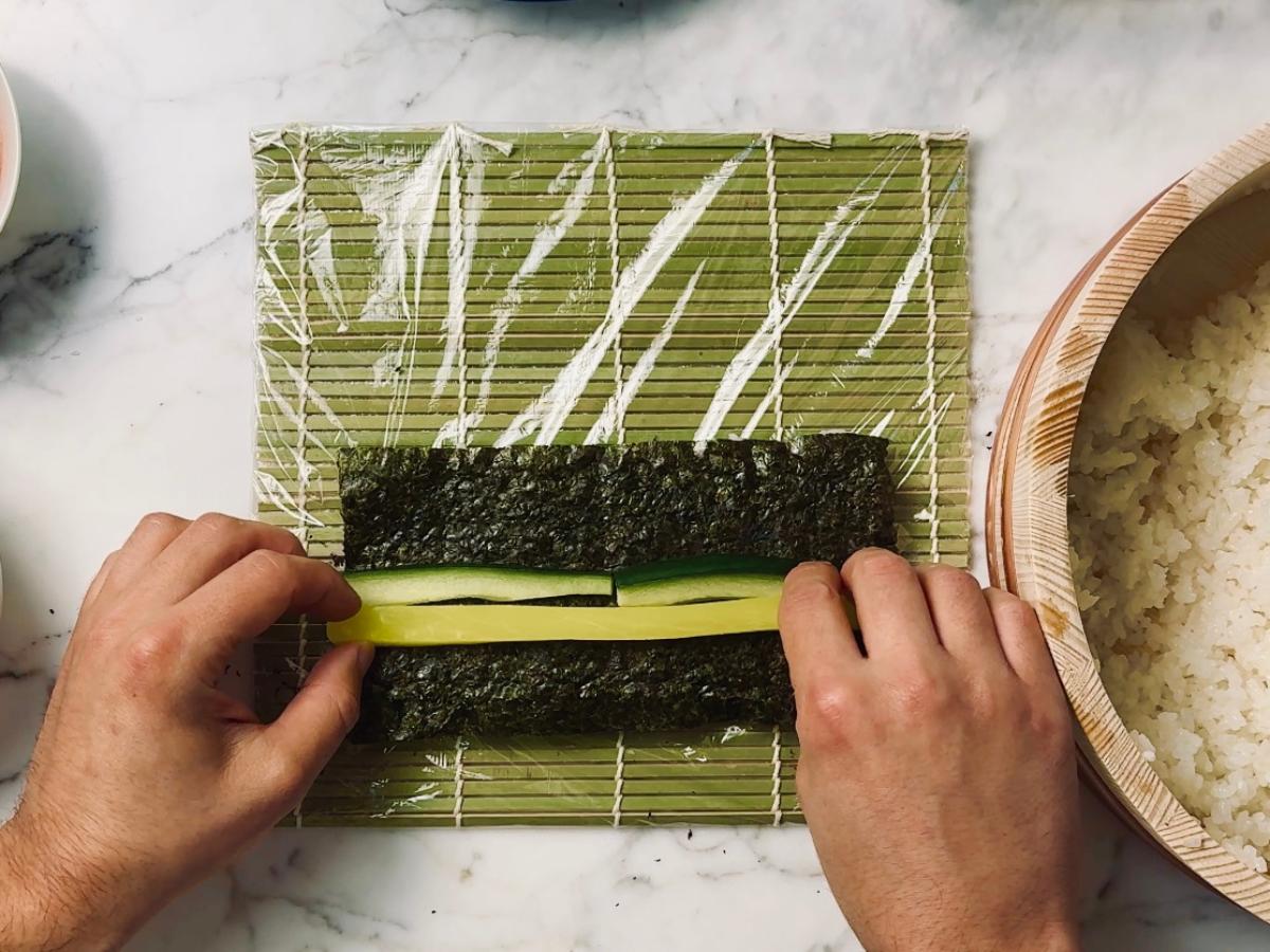 Sushi roll being assembled on a bamboo mat; nori seaweed is flipped to face upwards (on top of the rice), then strips of takuan and cucumber are placed on top.
