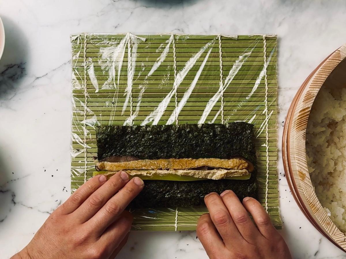Sushi roll being assembled on cling film laid over a bamboo sushi mat; sliced mushrooms and inari age are placed on the nori, leaving about one-third of the space clear at the top.