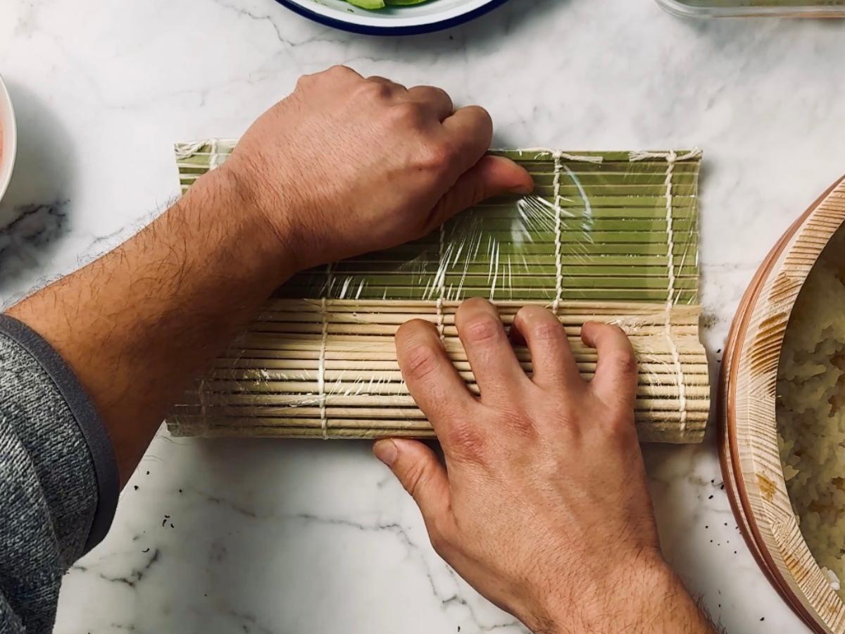 Two hands rolling a sushi roll with a bamboo mat wrapped in cling film; one hand holds the mat in place while the other pulls the top part away to tighten and seal the roll, keeping the fillings secure.