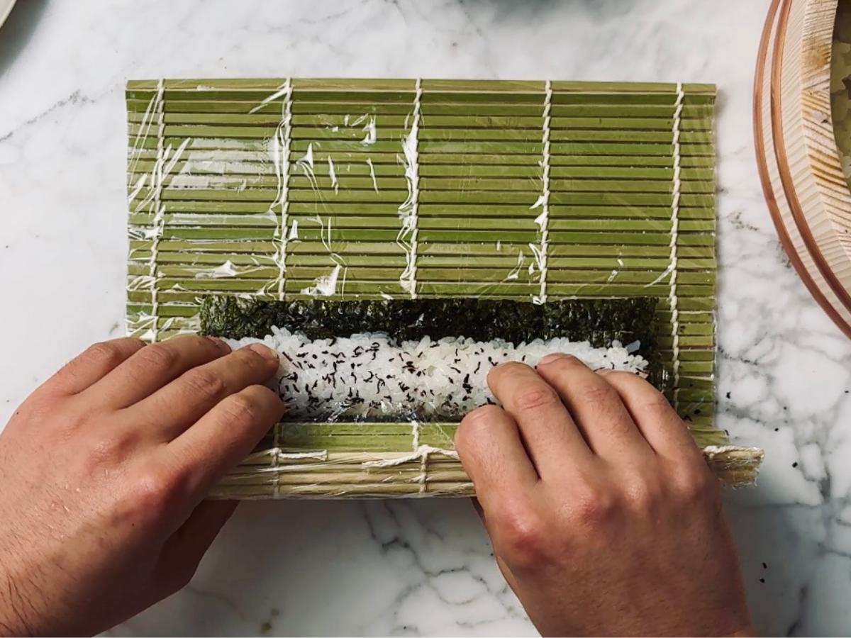 Two hands raising one end of a bamboo sushi mat to roll over sushi rice on nori seaweed.