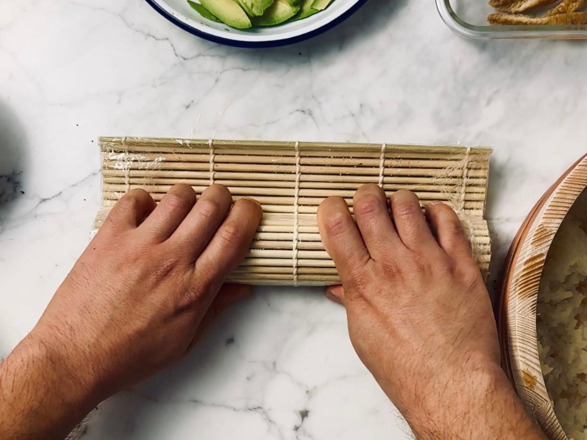 Two hands rolling a bamboo sushi mat with a sushi roll inside.