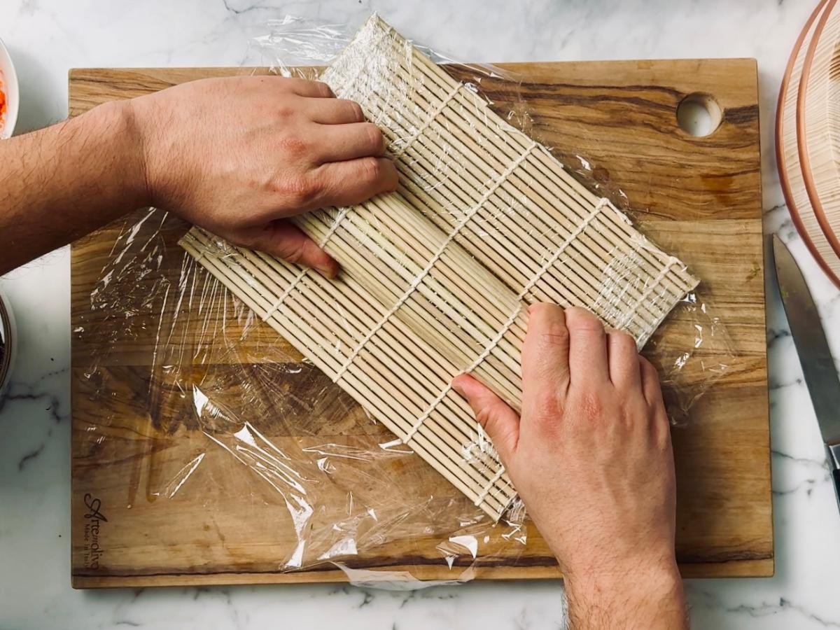 A roll of sushi covered in cling film on a wooden chopping board, with a bamboo mat over the top.