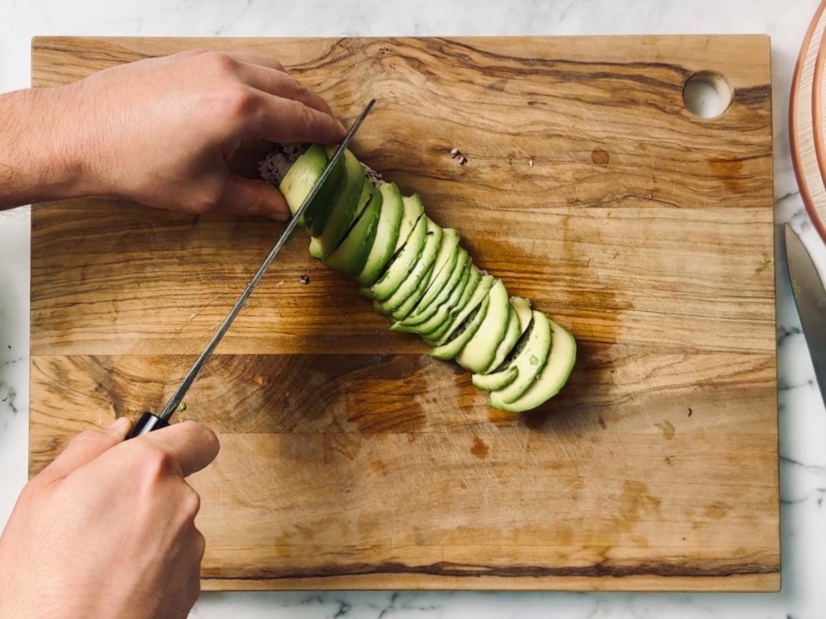 A pair of hands holding a large sharp knife, thinly slicing an avocado on a wooden chopping board.